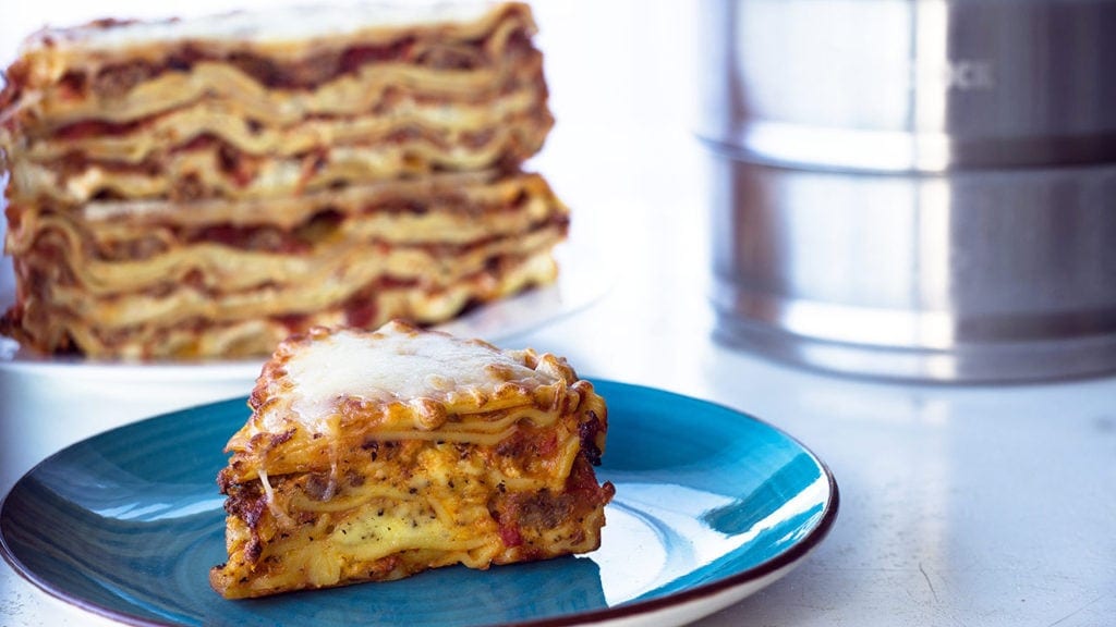 Lasagna slice sitting on a blue plate with the double decker lasagna in the background