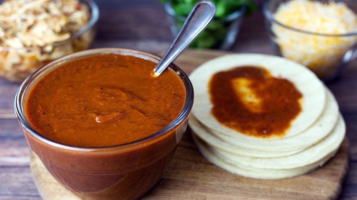 Large bowl of homemade enchilada sauce beside corn tortillas with ingredients for enchiladas in the back