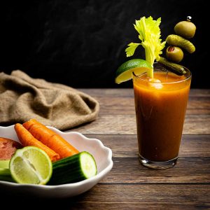 Bloody mary in a glass with a skewer of olives and pickles and a lime wedge with a celery stalk beside a white bowl with fresh veggies