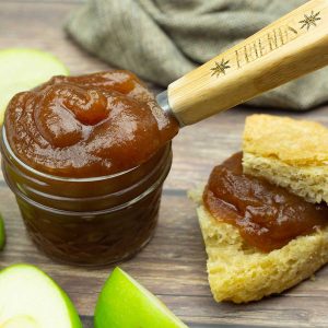 apple butter in a small jar next to sliced apples and some apple butter spread on crusty bread