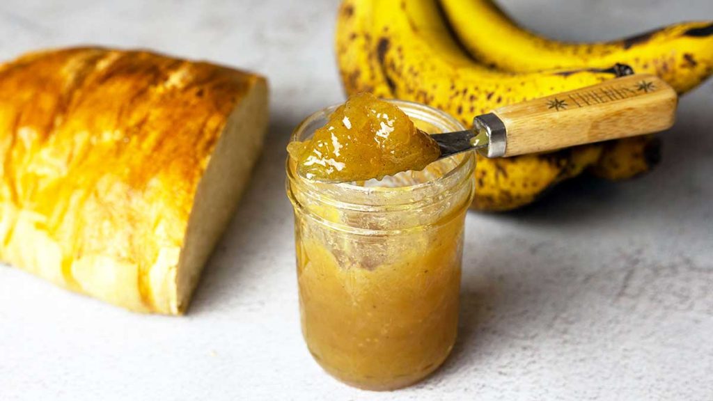 banana jam in a jar next to half a loaf of bread and a bunch of ripe bananas