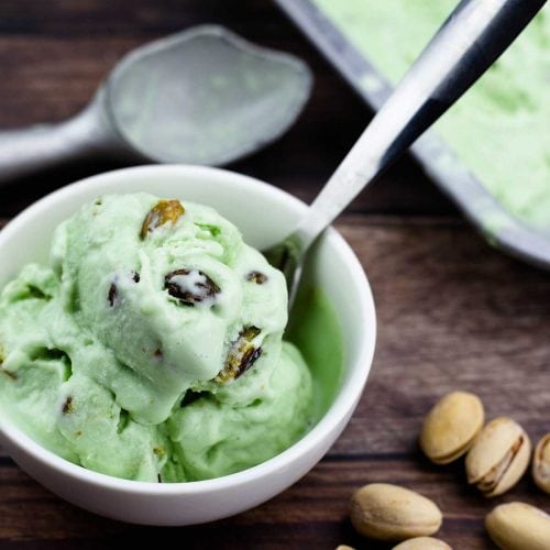 homemade pistachio ice cream in a bowl with pistachio nuts beside it an ice cream scoop behind it and the tray of ice cream to the right