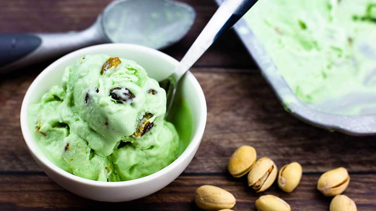 homemade pistachio ice cream in a bowl with pistachio nuts beside it an ice cream scoop behind it and the tray of ice cream to the right
