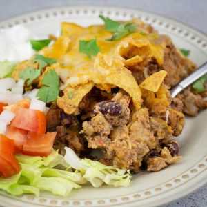 Walking taco casserole on a plate with lettuce, tomatoes, onions, and sour cream next to it