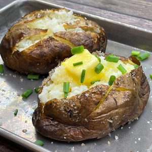 two baked potatoes on a tray one plain and one has butter and chives on it