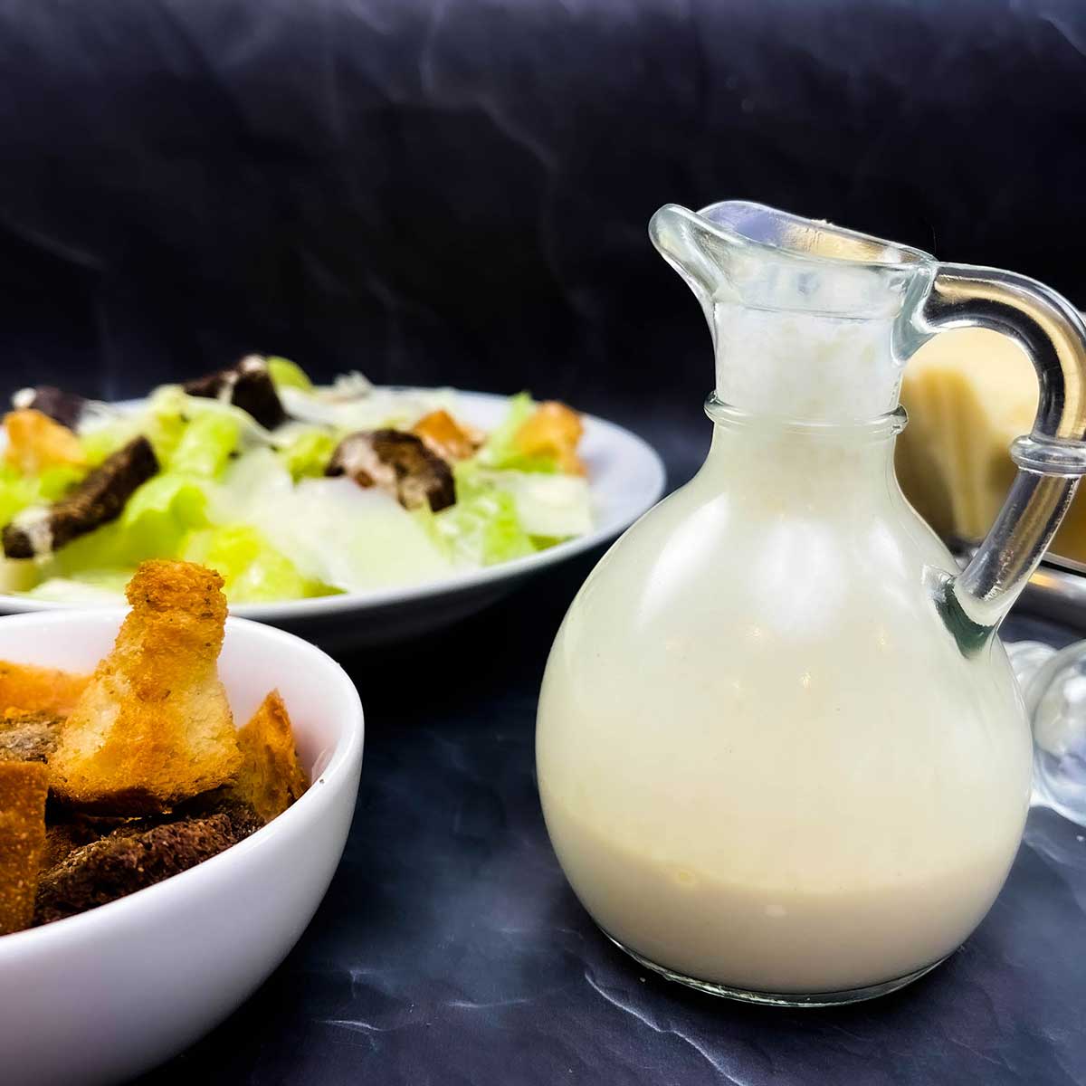 creamy caesar dressing in a glass container next to croutons and a caesar salad