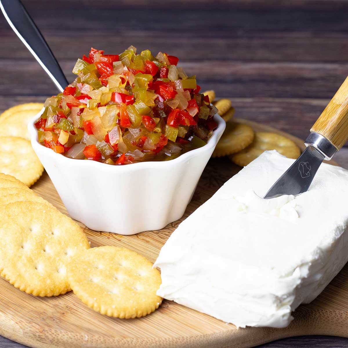 pepper and onion relish in a white bowl surrounded with cream cheese and crackers on a wood cutting board
