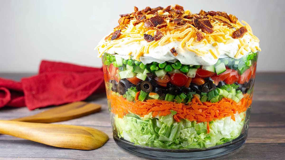 layered salad sitting on table with a red napkin and wooden serving utensils.