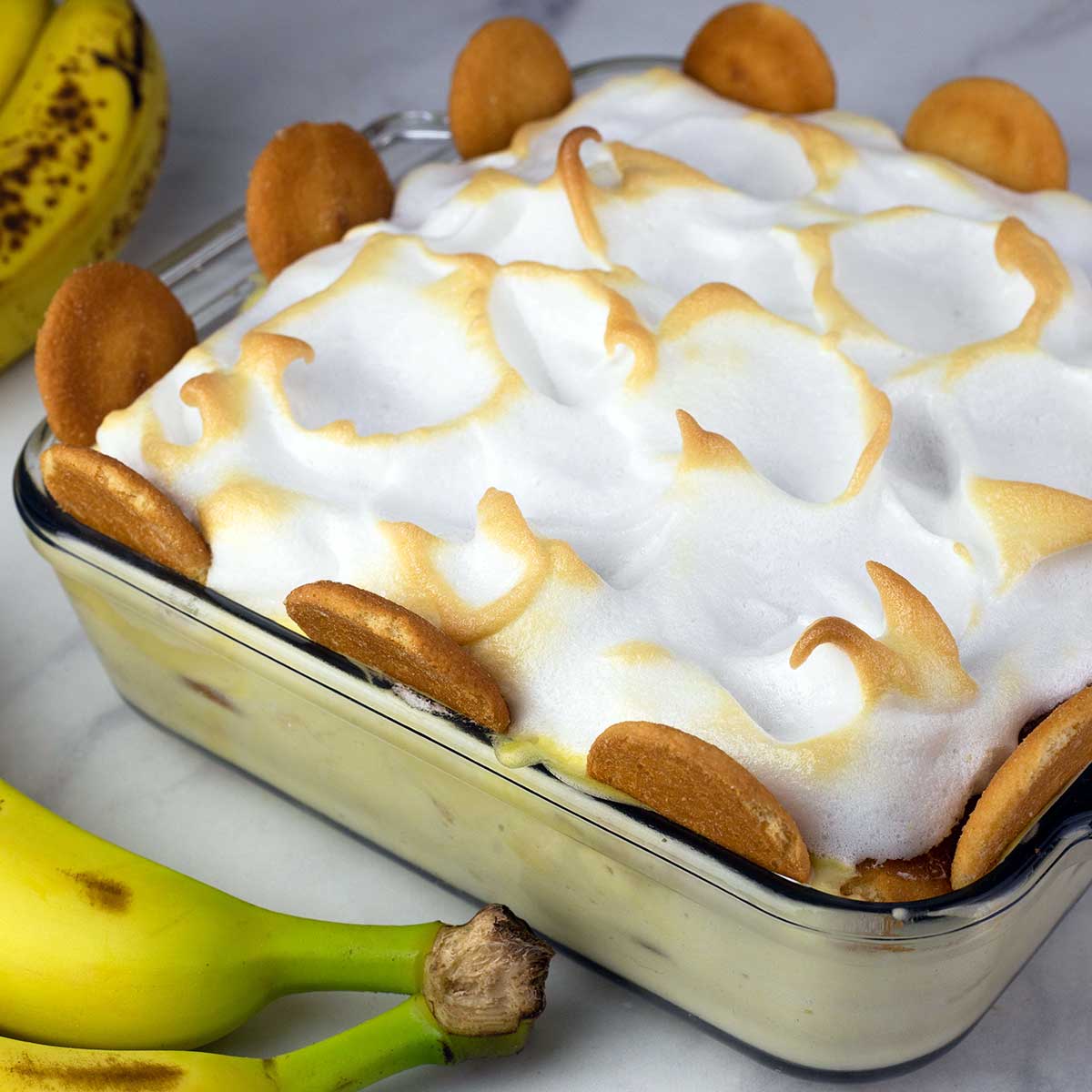 banana pudding with meringue topping in a glass square dish with cookies on the edge next to bananas