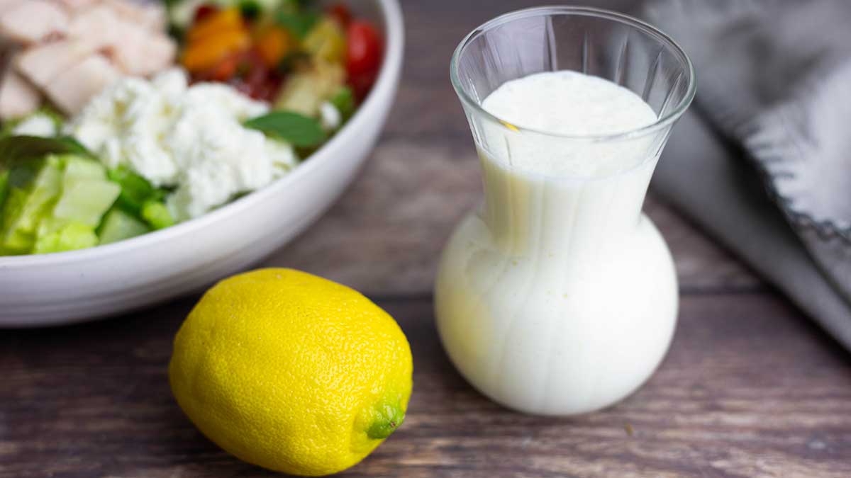 glass container with creamy lemon dressing next to a lemon and a salad.