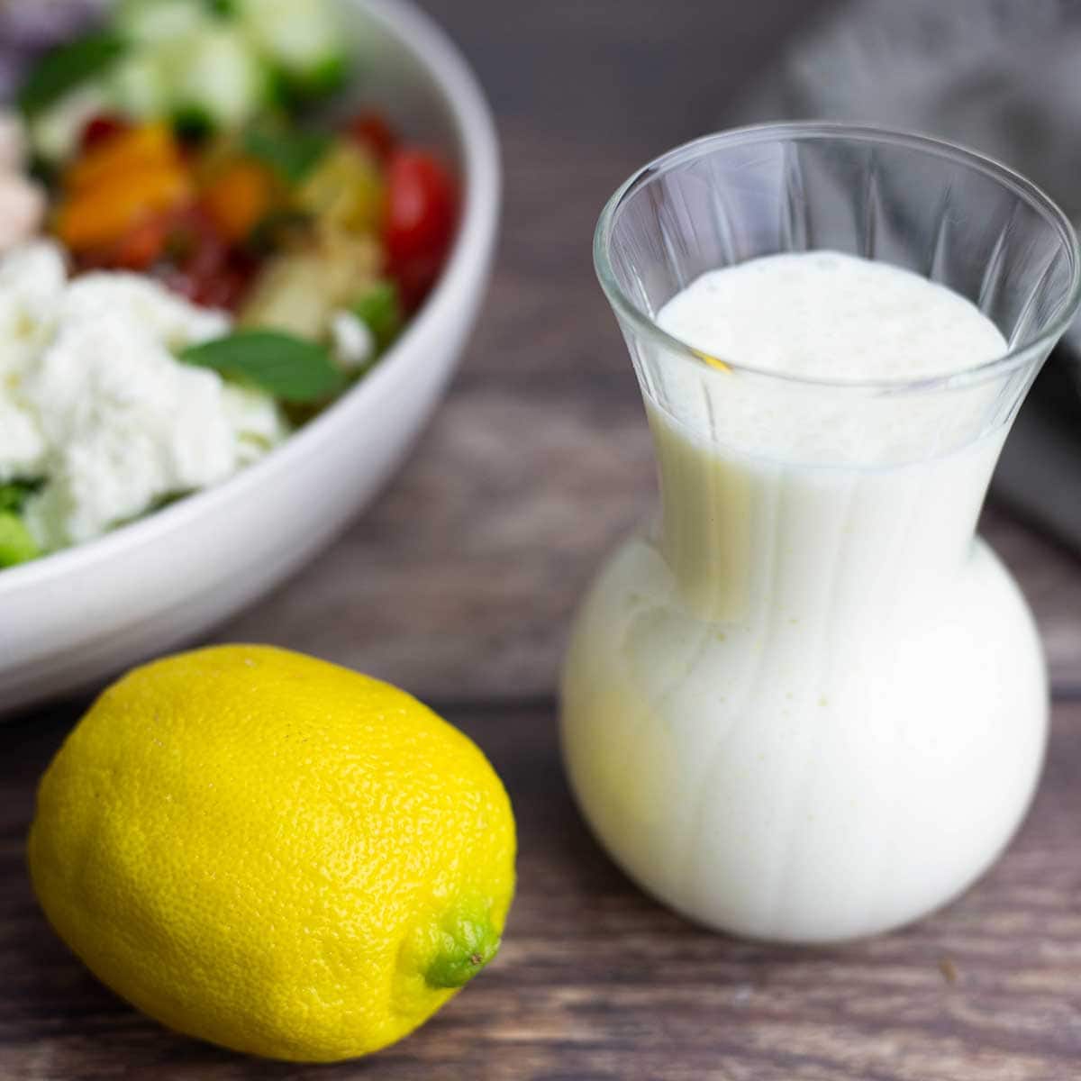 glass container with creamy lemon dressing next to a lemon and a salad.