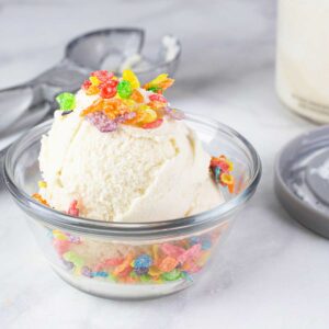 a bowl of fruity pebbles ice cream with fruity pebbles cereal on top and an ice cream container and scoop next to it.