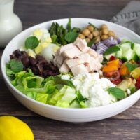 mediterranean salad in white bowl with a lemon and a bottle of lemon dressing beside it