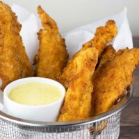 air fryer chicken strips in basket lined with parchment with a cup of honey mustard sauce.