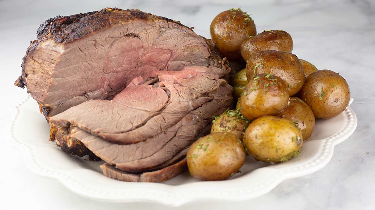 air fryer leg of lamb sliced on a white platter next to potatoes.