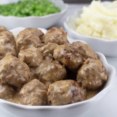 Swedish Meatballs in gravy in a white serving bowl next to mashed potatoes and peas.