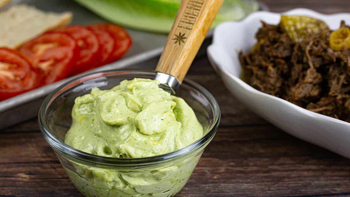 Homemade avocado oil ranch flavored mayonnaise in a clear glass bowl beside the fixings for a pot roast sub sandwich. 