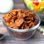 Homemade bacon bits in a small glass bowl in front of a salad and dressing.