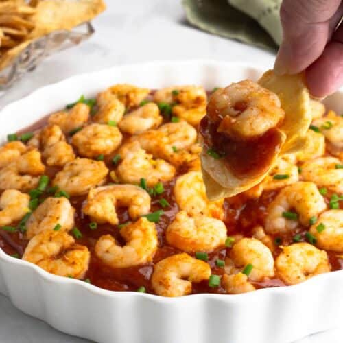 White serving dish with cold shrimp dip and a hand with a cracker is scooping out some of the dip.