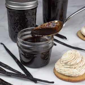 Homemade Vanilla Bean Paste in a small jar with spoon showing the thickness of it with a cookie topped with whipped cream made with vanilla paste beside it.