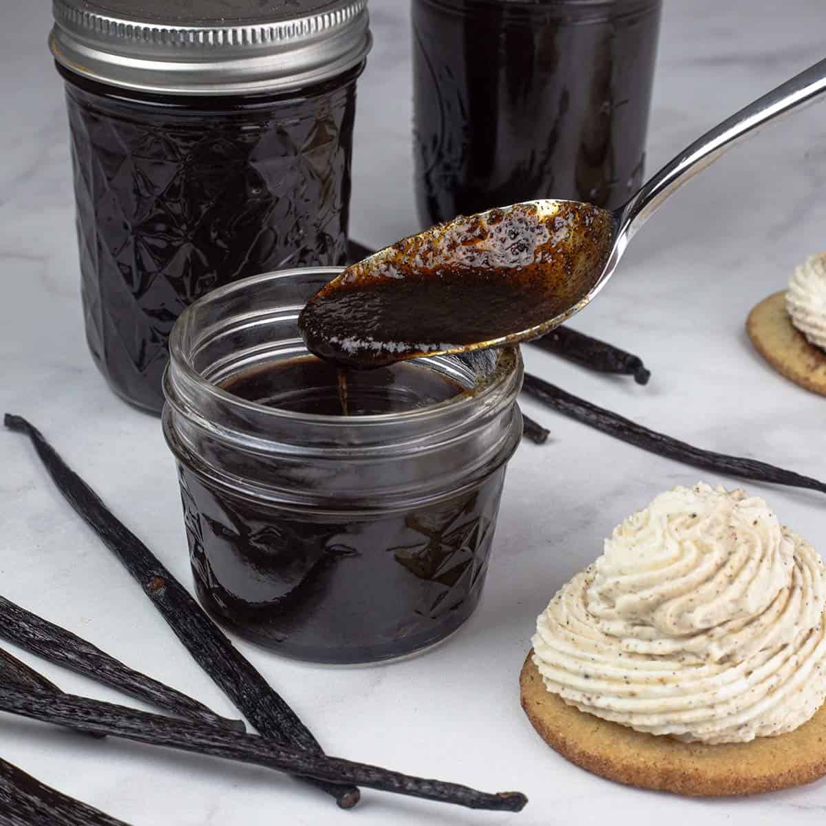 Homemade Vanilla Bean Paste in a small jar with spoon showing the thickness of it with a cookie topped with whipped cream made with vanilla paste beside it.