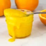 Homemade orange curd in a jar with a spoon and cut and whole oranges behind it.