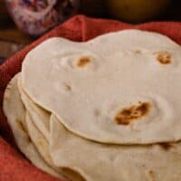 Homemade flour tortillas stacked in a basket lined with a red towel.