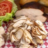 Smoked Turkey Breast sliced thin on a red and white checkerboard deli paper with the whole turkey roast behind it and bread, lettuce, tomato slices to the right.