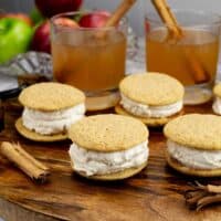 Apple Cider Whoopie pies on a wood tray with apple cider in glasses and cinnamon sticks.