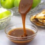 homemade caramel sauce dripping off of a spoon into a bowl with apple slices and an apple strudel behind it.