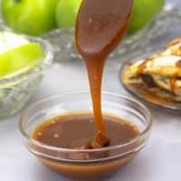 homemade caramel sauce dripping off of a spoon into a bowl with apple slices and an apple strudel behind it.