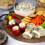 cold spinach and artichoke dip in a bowl surrounded by fresh vegetables, stuffed tomatoes, and pinwheel sandwiches.