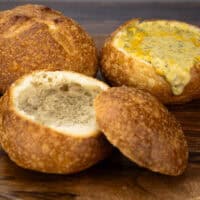 three homemade bread bowls on a wood platter. One is whole, one is hollowed out and one is filled with cheddar broccoli soup.