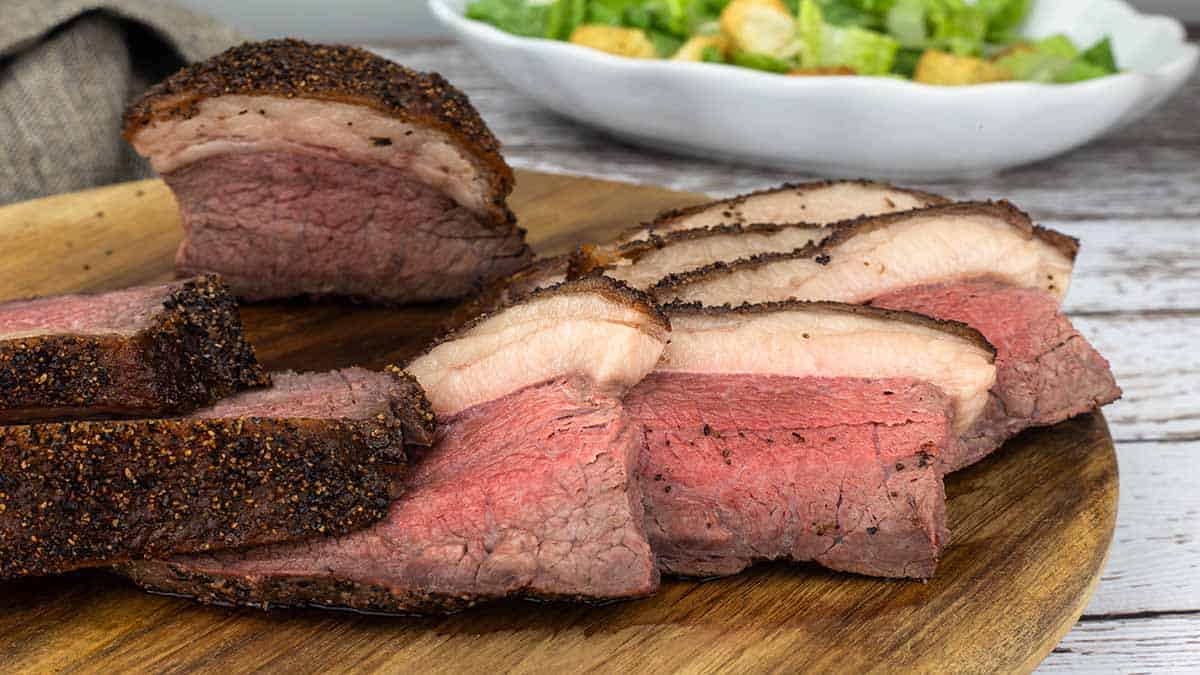 Sliced Rare Picanha roast on a wood cutting board with a salad behind it. 