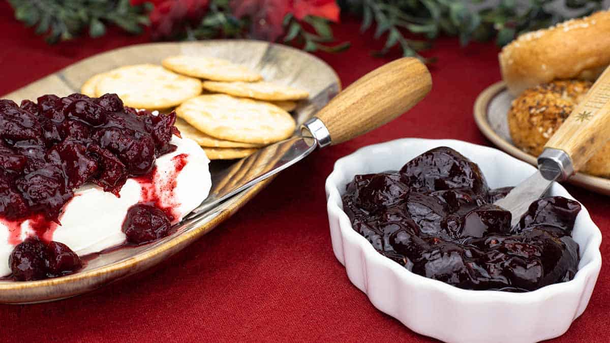 Cranberry Jelly in a white bowl next to bagels and cranberry preserves on top of cream cheese with crackers on a platter. 