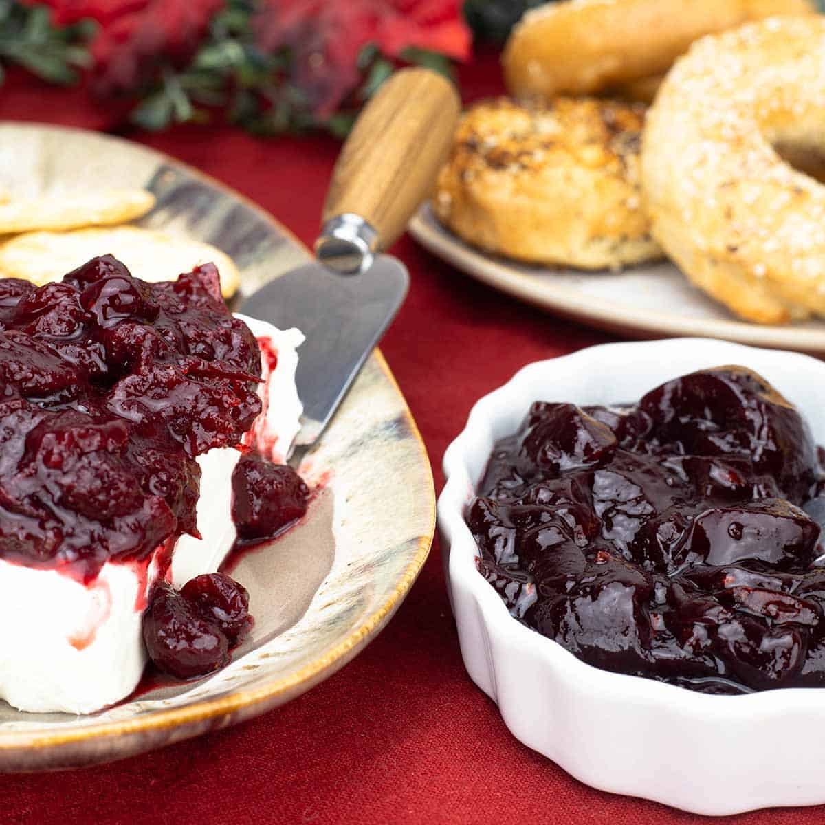 Cranberry Jelly in a white bowl next to bagels and cranberry preserves on top of cream cheese with crackers on a platter.