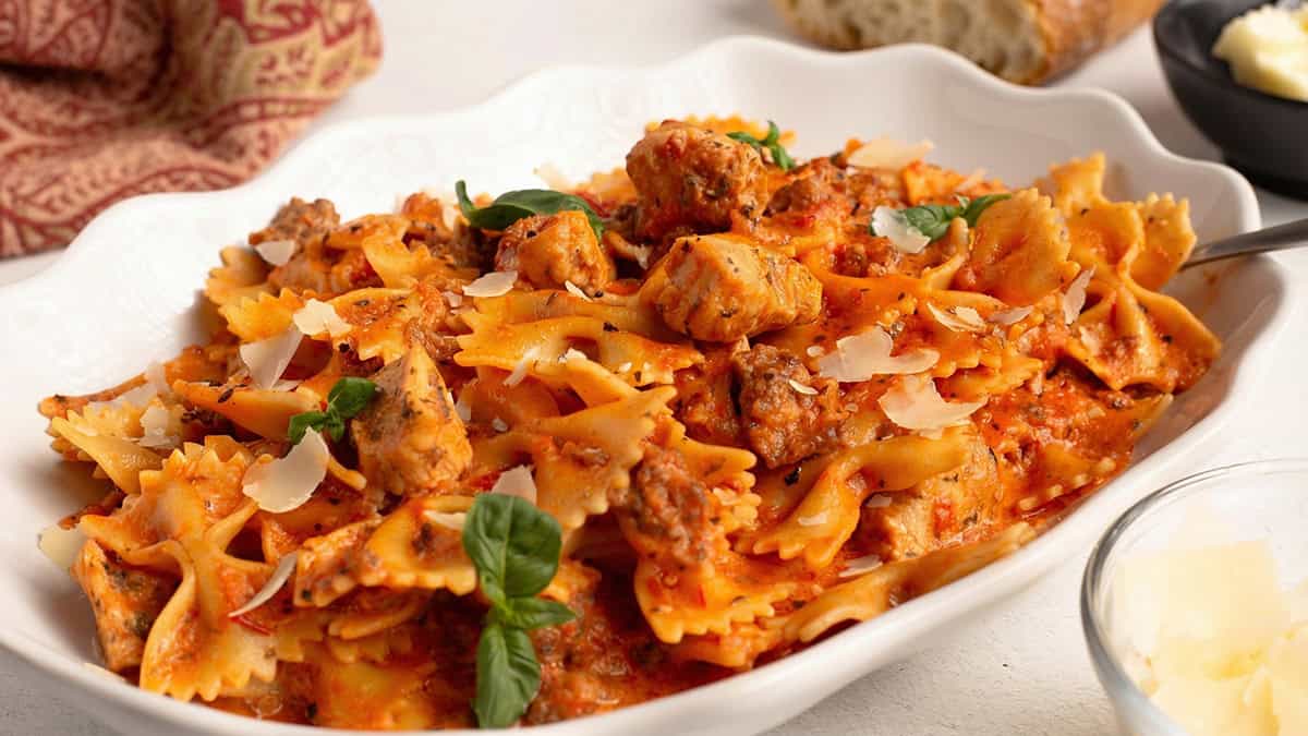 Parma Rosa Pasta with Chicken and Sausage served in a white bowl, showing creamy pink tomato sauce coating bow tie pasta with Italian sausage and chicken, topped with shaved parmesan and fresh basil.