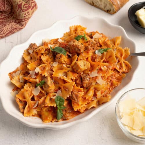 Square crop of Parma Rosa Pasta with Chicken and Sausage in a white bowl with creamy pink sauce, bow tie pasta, sausage, and chicken garnished with parmesan and basil.