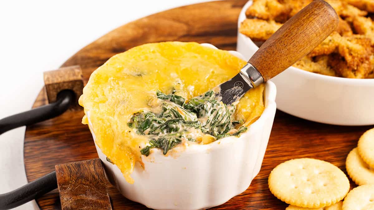Cheesy spinach dip ina white bowl with crackers and pasta chips beside it.