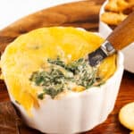 Cheesy spinach dip ina white bowl with crackers and pasta chips beside it.
