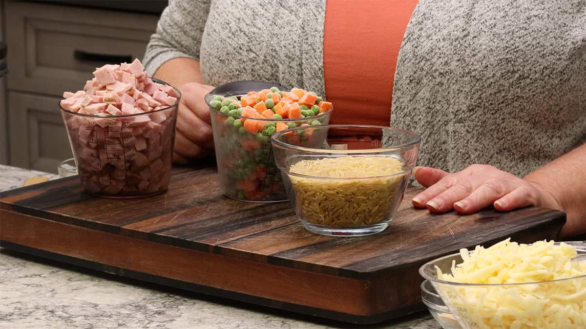 Bowls of diced ham frozen peas and carrots orzo pasta and shredded cheese on a cutting board