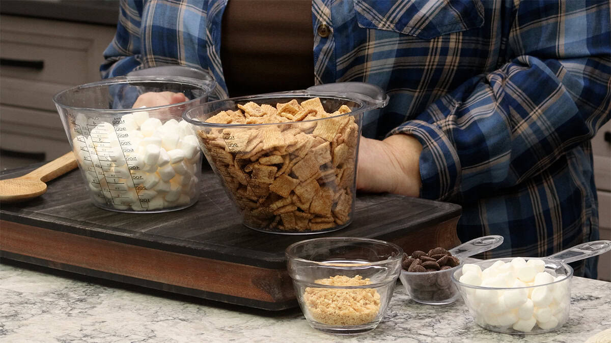 Ingredients for Cinnamon Toast Crunch bars laid out on a countertop: a box of Cinnamon Toast Crunch cereal, a bowl of cereal, marshmallows in a glass bowl, butter, and vanilla extract