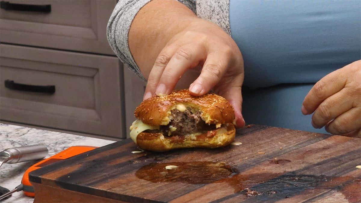 Hands holding a juicy air fryer burger with a bite taken out of it on a wooden cutting board, showing the cooked interior with melted cheese and sauce.
