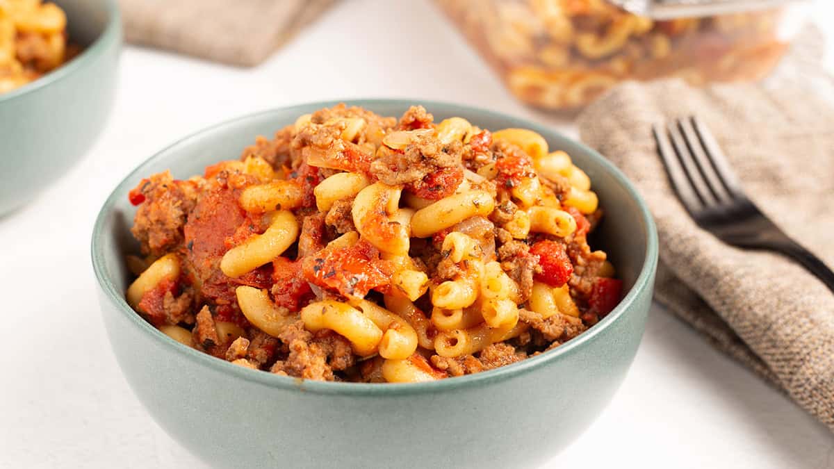 Close-up hero shot of American goulash in a teal ceramic bowl with elbow macaroni, ground beef, and rich tomato sauce, a second bowl and glass baking dish visible in the background.