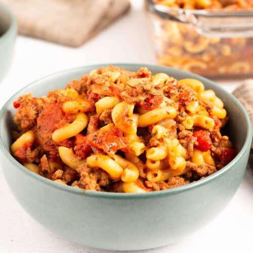 Square image of a teal bowl filled with American goulash showing elbow macaroni mixed with seasoned ground beef and tomato sauce, with a glass dish of extra goulash in the background.