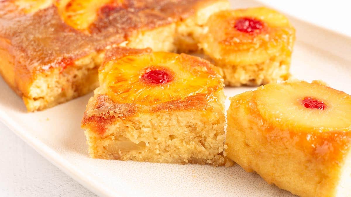 Squares of pineapple upside down cake arranged on a cream platter, with a caramelized pineapple ring and maraschino cherry on top of each piece.