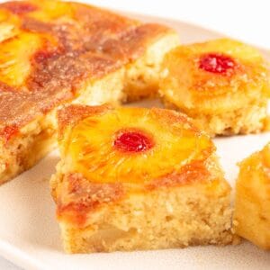 Close up of pineapple upside down cake squares topped with caramelized pineapple rings and maraschino cherries on a cream platter.