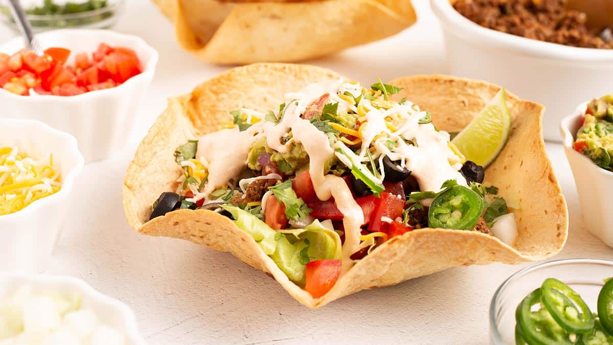 Crispy air fryer flour tortilla taco salad bowl filled with seasoned ground beef, shredded lettuce, diced tomatoes, shredded cheese, and a drizzle of crema dressing