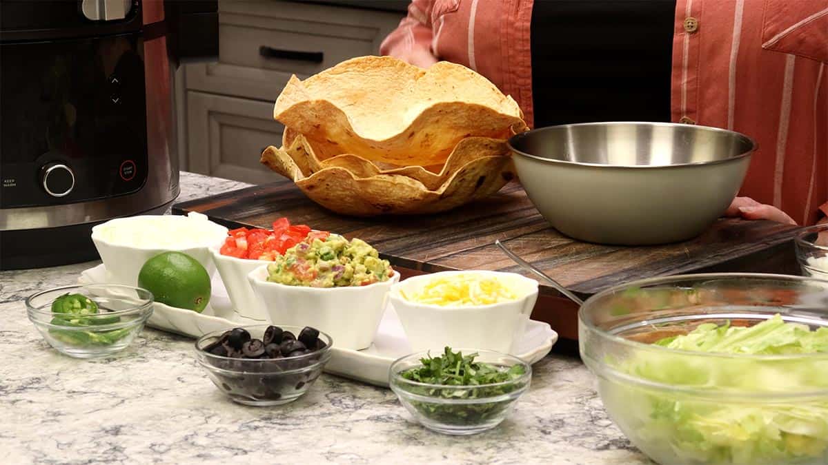 Counter setup with stacked crispy tortilla bowls, a stainless mixing bowl, and small dishes of taco salad toppings including ground beef, refried beans, lettuce, tomatoes, cheese, olives, and crema