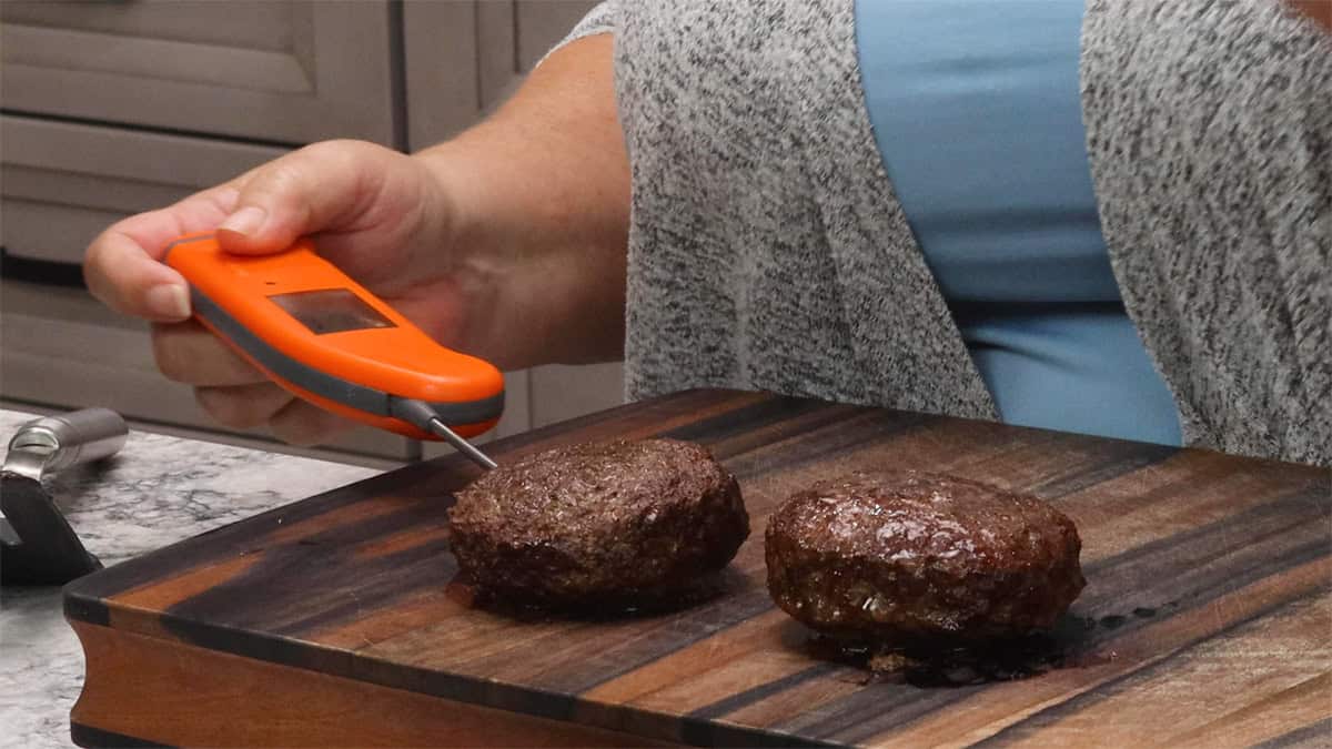 Checking the internal temperature of two cooked air fryer burger patties on a wooden cutting board using an orange instant-read thermometer.
